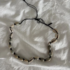 Snäckhalsband med svart snöre - Unikt halsband med naturliga snäckor i beige och grå toner, trädda på ett svart snöre med knytfunktion. Perfekt för dig som gillar strandvibbar och boho-stil. Snäckorna är ovala och ger ett coolt, avslappnat uttryck.