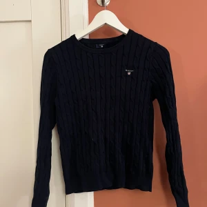 Marinblå kabelstickad tröja GANT - Snygg marinblå kabelstickad tröja från GANT med rund hals och broderad logga på bröstet. Tröjan har lång ärm och ribbade muddar vid hals, ärmslut och nederkant. Inte använd på ett långt tag så med en tvätt blir den som ny! Storlek 158/164 så xs/s. Priset kan diskuteras!🤗