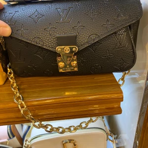 Micky Svart handväska  - Snygg svart handväska med  guldfärgad kedja. Väskan har en elegant låsdetalj i guld och är tillverkad i präglat skinn. Perfekt storlek för det viktigaste och en riktigt lyxig vibe.