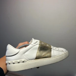 Valentino Garavani sneakers i vitt och guld - Säljer ett par snygga Valentino Garavani sneakers i vitt skinn med metallicguld detalj över snörningen. Skorna har platt sula, rund tå och coola nitar baktill. Perfekt för dig som gillar lyxiga och trendiga sneakers med unik design.