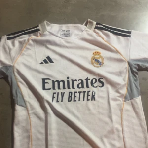 Real Madrid hemmatröja Adidas vit - Snygg Real Madrid hemmatröja från Adidas i vitt med mörkblå detaljer och klubbmärke på bröstet. Tröjan har korta ärmar, rund hals och gula inslag längs sidorna. Tillverkad i lätt och ventilerande material, perfekt för fotboll eller supporterliv.