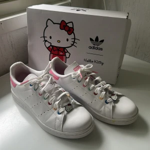 Adidas Stan Smith Hello Kitty sneakers - (SKICKAS NYTVÄTTADE) Unika Adidas Stan Smith Hello Kitty sneakers i vitt skinn med rosa detaljer och färgglada blommor på snörningen. Skorna har Hello Kitty-motiv på tungan och hälen samt charm på snörena. Perfekt för dig som älskar söta och lekfulla detaljer.