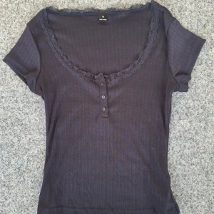 Svart ribbad topp med spets XS - Säljer en svart ribbad topp i storlek XS med korta ärmar och snygg spetskant runt den djupa halsringningen. Toppen har tre knappar framtill och är gjord i mjukt material som sitter skönt mot kroppen. Perfekt för dig som gillar feminina detaljer och enkel stil.