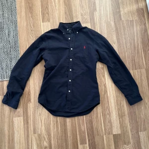 Svart skjorta från Ralph Lauren S - Klassisk mörkblå skjorta från Ralph Lauren i slim fit med lång ärm och knappar framtill. Skjortan har röd broderad logga på bröstet och button down-krage. Tillverkad i mjuk bomull, perfekt för en clean och stilren look.
