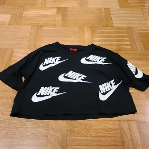 Svart croppad Nike topp XS - Svart croppad topp från Nike i storlek XS. T-shirten har korta ärmar och är täckt med stora vita Nike-loggor både fram och bak. Tillverkad i mjuk bomull, perfekt för dig som gillar sportig streetstyle och vill sticka ut med en bold look.