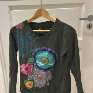 Grön långärmad tröja Desigual - Unik grön långärmad tröja från Desigual med färgglada blommiga och cirkulära applikationer i rosa, turkos, lila och gult. V-ringad hals och coolt broderade detaljer som ger en kreativ vibe. Perfekt för dig som gillar att sticka ut med din stil.