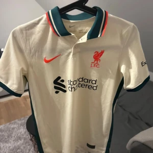 Liverpool Nike fotbollströja Dri-FIT L - Liverpool FC:s officiella bortatröja från Nike i Dri-FIT-material. Tröjan är beige med gröna detaljer på krage och ärmslut, röda accenter och klubbmärke på bröstet. Klassisk pikékrage och tryckta sponsorer framtill. Perfekt för dig som älskar fotboll och vill sticka ut.