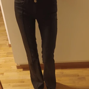 Svarta bootcut-byxor från Nelly - Snygga svarta byxor från Nelly med bootcut-snitt och två dekorativa knappar i midjan. Använda 1 gång. Ser ut som skinnbyxor men det är liksom inte skinnmaterial.