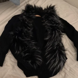 Svart kofta med pälsdetaljer Novo Style - Svart kofta från Novo Style i storlek 10 med fluffiga pälsdetaljer längs kragen och framstycket. Koftan har långärmad design och ribbad struktur i materialet, vilket ger en cool och unik look. Perfekt för dig som vill sticka ut med en statement piece.