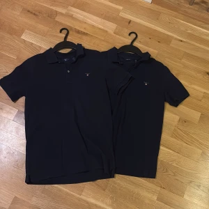 Marinblå pikétröja från GANT - Två stilrena marinblå pikétröjor från GANT med klassisk krage och knappslå. Tröjorna har broderad GANT-logga med sköld på bröstet och är tillverkade i mjuk bomull. Perfekt för dig som gillar en clean och tidlös look. Säljer för sig därför två annonser