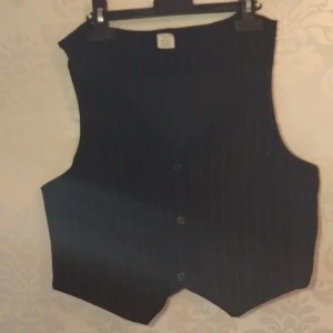 Svart randig väst från Lindex - Klassisk svart väst från Lindex med tunna vita ränder och knäppning framtill. Snyggt skräddad modell med spetsig nederkant och diskreta knappar. Perfekt för dig som gillar en stilren och tidlös look.