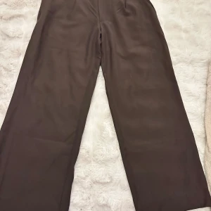 Bruna kostymbyxor från 157, storlek M - Snygga bruna kostymbyxor från 157 i storlek M. Byxorna har rak och vid passform, hög midja och klassisk dragkedja med knappstängning. Materialet är mjukt och har en lätt glansig finish som ger en clean look.