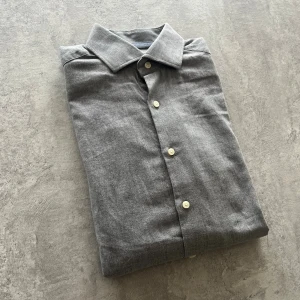 Grå skjorta från Riley slim fit - Stilren grå skjorta från Riley i slim fit-modell. Skjortan har klassisk krage, vita knappar och långa ärmar. Tillverkad i mjuk bomull som känns skön mot huden. Perfekt för dig som gillar en clean och enkel look.