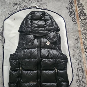 Moncler Väst - Snygg svart dunväst från Moncler med avtagbar huva och två dragkedjeförsedda fickor framtill. Västen har quiltad design, Moncler-logga på bröstet och coolt serietryck på insidan. Perfekt för lager-på-lager och streetstyle. 