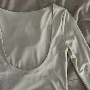 Vit långärmad topp med djup urringning backtill - Säljer en stilren vit långärmad topp med djup, rundad urringning backtill. Toppen har en slät och mjuk yta, vilket ger en clean look. Passformen är normal och materialet känns lätt och stretchigt, perfekt för att styla med jeans eller kjol.