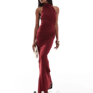 Vinröd långklänning med öppen rygg - Supersnygg vinröd långklänning med halterneck och öppen rygg. Klänningen har en figurnära passform, slits på sidan och draperade detaljer längs midjan. Tillverkad i ett glansigt, stretchigt material som ger en lyxig känsla.