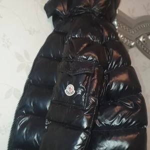 Svart pufferjacka från Moncler - Säljer en svart, glansig pufferjacka från Moncler med klassisk logga på ärmen och dragkedja framtill. Jackan har en stor huva och rejäla fickor, perfekt för kalla dagar. Den har en oversized look och är riktigt varm och skön.ÄKTa!