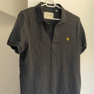 Mörkgrå pikétröja från Lyle & Scott - Snygg mörkgrå pikétröja från Lyle & Scott med klassisk krage och knappslå. Tröjan har korta ärmar och är gjord i mjuk bomull. På bröstet finns den ikoniska gula fågeln broderad. Perfekt för en chill och stilren look.