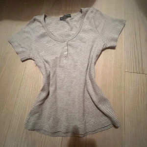 Randig topp från Brandy Melville - Superfin topp från Brandy Melville i beige och vit med smala ränder. Modellen har korta ärmar, rundad halsringning och tre knappar framtill. Mjuk och stretchig bomull som sitter snyggt och är perfekt för en chill look.