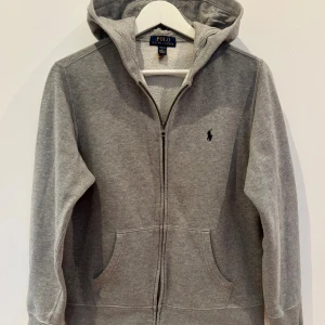Polo Ralph Lauren GRÅ FLEECE ZIP HOODIE - Ljusgrå zip-hoodie från Polo Ralph Lauren. Silverfärgad dragkedja och sidofickor.  Storlek L (12-14 år).  Inköpt på Kids Brand Store i september förra året för    1 195 kr. Mycket fint skick.  Hoodien är nytvättad utan sköljmedel.  