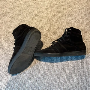 Svarta Saint Laurent high top sneakers - Svarta high top sneakers från Saint Laurent Paris i lyxig mocka och skinn. Klassisk siluett med snörning, rund tå och diskreta detaljer. Perfekta för dig som vill ha en clean och stilren look med exklusiv känsla. Skorna är köpte för 4 år sedan i Saint lorant butiken i Paris. Skorna är sedan dess helt oanvända och har inte en enda skråma eller någon annan slitning. Skorna är alltså i helt nytt skick och har stått näst in till orörda sen dagen dom köptes.
