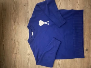 Blå stickad tröja från Ami Paris (trade bara) - Blå stickad tröja från Ami Paris med vitt hjärta och bokstaven A på bröstet. Tröjan har rund hals, långa ärmar och breda ribbade muddar vid ärmslut och nederkant. Materialet är mjuk bomull och passformen är normal. Perfekt för dig som gillar stilrena detaljer.