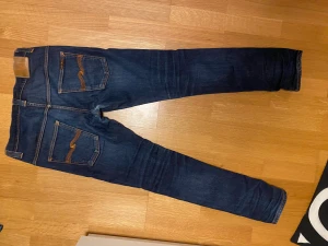 Mörkblå skinny jeans från Nudie - Säljer ett par mörkblå skinny jeans från Nudie med klassisk orange söm och snygga bakfickor med vågig brodyr. Har storlek 32/32 men sitter mer som 30/32 Perfekt passform för dig som har lite smalare ben. Obs! Det är ett litet hål som syns på bild 4. Går att lämna in gratis i nudie jeans butiken för reparation. Kontakta gärna vid frågor!
