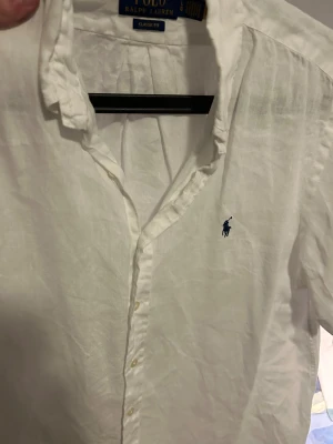 Vit linneskjorta från Polo Ralph Lauren - Vit klassisk skjorta från Polo Ralph Lauren i storlek L med korta armar. Tillverkad i linne med klassisk passform och knappar framtill. Har den ikoniska blå Polo-loggan broderad på bröstet och en snygg krage. Perfekt för en clean och fräsch look.