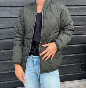 Olivgrön quiltad bomberjacka NY Jack&Jones - Snygg olivgrön bomberjacka med quiltad design och dragkedja framtill. Jackan har ribbade muddar vid hals och ärmslut samt två sidofickor. Perfekt för dig som vill ha en stilren och trendig look under höst och vinter.