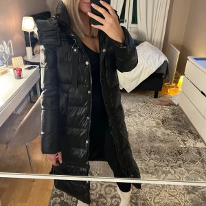 Vinterjacka - Säljer en svart, lång pufferjacka med huva och snygga detaljer som dragkedja och knappar. Perfekt för kalla dagar och har en modern, oversized look.