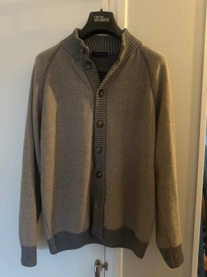 Grå cardigan från italienska Catapano - Snygg grå cardigan från Catapano med ribbad krage och muddar. Koftan har knäppning med fem knappar framtill och är tillverkad i en mjuk ullblandning. Perfekt för kyliga dagar och har en clean, klassisk look. Köpt i Italien och den är gjord i Italien. Tyvärr aningen för stor för mig. Skulle uppskatta till en storlek M men kolla måtten på bilderna för säkerhets skull!