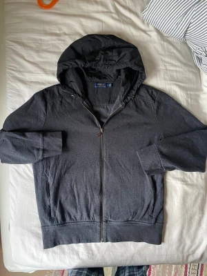 Polo Ralph Lauren cardigan - Grå cardigan från Polo Ralph Lauren med huva och snörning. Jackan har dragkedja framtill och på fickorna, samt detaljer i syntetmaterial på axlar och huva. Stilren design med diskret logga på bröstet. Perfekt för dig som gillar sportig och clean look. Skicket är väldigt bra, utan defekter. Kontakta mig om frågor! 😊