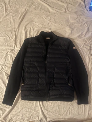 Svart Moncler Jacka  - Snygg svart pufferjacka från Moncler med quiltad front och ärmar i stickad ull. Jackan har två dragkedjefickor, hel dragkedja framtill och klassisk Moncler-logga på ärmen. Perfekt för dig som vill ha en stilren och exklusiv look.