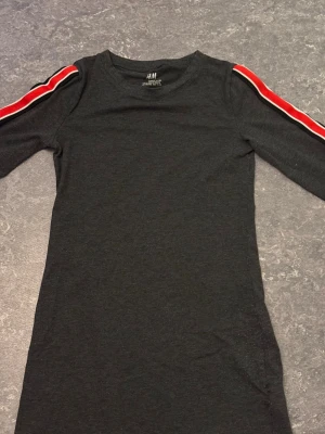 Svart långärmad topp med ränder H&M - Svart långärmad topp från H&M med coola röda och vita ränder längs axlarna. Rund halsringning och slim passform. Perfekt för dig som gillar sportig stil och vill ha något enkelt men ändå med en snygg detalj på ärmarna.