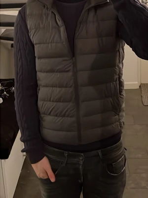 Grå dunväst från Uniqlo Ultra Light Down - Säljer en grå dunväst från Uniqlo Ultra Light Down. Västen har en quiltad design med horisontella sömmar, hög krage och dragkedja framtill. Perfekt att slänga över en hoodie eller tröja för extra värme. Lätt och smidig att bära, passar till många olika stilar.