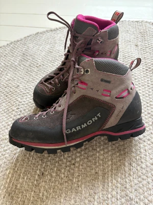Garmont vandringskängor grå/rosa - Säljer ett par robusta vandringskängor från Garmont i grått och mörkrosa med detaljer i rosa och svart. Skorna har grov Vibram-sula, snörning och är tillverkade i mocka och syntet. Perfekt för dig som gillar äventyr och vill ha snygga kängor med coola färgdetaljer.