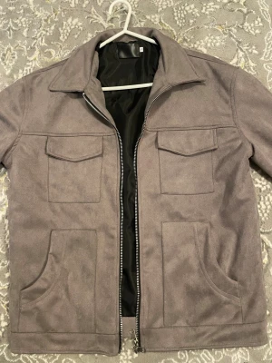 Grå mocka overshirt med fickor - Snygg grå overshirt i mocka-look med fyra stora fickor framtill och silverfärgad dragkedja. Jackan har klassisk krage, långa ärmar med knapp vid ärmslut och svart innerfoder. Perfekt för lager-på-lager och chill stil.