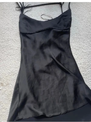 Svart satin kortklänning - Snygg svart kortklänning i satin med smala axelband och lätt v-ringad hals. Klänningen har en slät, glansig yta och sitter figurnära upptill med lite lösare passform nedtill. Perfekt för dig som gillar minimalistisk och stilren look.