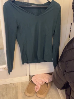 Mörkgrön långärmad v-ringad topp - Snygg mörkgrön långärmad topp med v-ringning och slim passform. Enkel och stilren design som funkar till många olika looks. Mjukt material som känns skönt mot huden och sitter snyggt på kroppen.