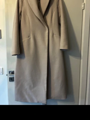 Beige kappa med bälte från Ellos - Säljer en stilren beige kappa från Ellos med bred krage och bälte i midjan. Kappan har långärmad design, slät yta och två diskreta fickor framtill. Perfekt för dig som gillar klassisk look och vill ha något som håller värmen under kyliga dagar.