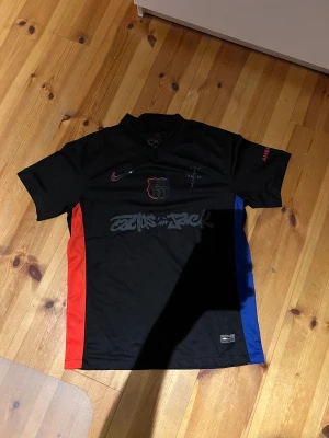 Svart Nike x FC Barcelona Cactus Jack tröja - Limited edition FC Barcelona x Cactus Jack tröja från Nike i svart med röda och blå sidopaneler. Snyggt tryck på bröstet, klubbmärke och Nike-logga i rött/blått. Materialet är 100% polyester, perfekt för fotboll eller streetwear. UNHCR-tryck på ryggen och coola detaljer på ärmarna.