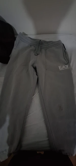 Grå mjukisbyxor EA7 Emporio Armani - Grå mjukisbyxor från EA7 Emporio Armani med logga tryckt på vänster lår. Byxorna har snörning i midjan och ribbade muddar vid bensluten. Sköna och stilrena, perfekta för chill eller träning. Materialet är mjukt och känns riktigt bekvämt.