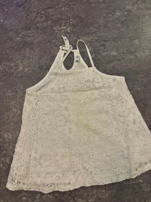 Vit spetsig linnetopp med knyt - Supersöt vit linnetopp med blommig spets och knyt i nacken. Toppen är ärmlös och har en lätt A-linjeformad siluett. Perfekt för varma dagar och ger en romantisk vibe med sitt detaljerade spetsmönster.