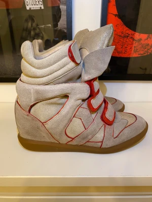 Isabel Marant Étoile höga sneakers - Säljer ett par höga sneakers från Isabel Marant Étoile i beige mocka med röda detaljer och kardborreband. Skorna har en chunky siluett, platt gummisula och rund tå. Perfekt för dig som gillar statement-skor med cool design och extra komfort.