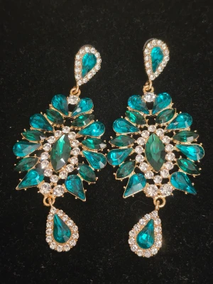 Strass örhängen  - Bilden visar ett par glittriga örhängen med turkosa och klara stenar, infattade i guldfärgad metall. Ingen väst syns på bilden.