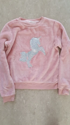 Tröja - Supersöt rosa tröja från KappAhl i fluffigt material. Framtill finns en stor enhörning i glittriga silverpaljetter som verkligen poppar. Tröjan har långa ärmar och mjuka muddar vid ärmslut och nederkant. Perfekt för dig som älskar magiska detaljer! Några små noppror.