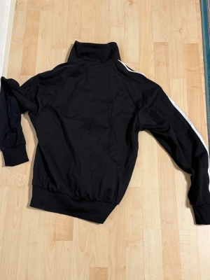 Adidas Y-3 svart track jacket L - Svart track jacket från Adidas Y-3 Yohji Yamamoto med vita ränder längs ärmarna och Y-3 logga på bröstet. Jackan har hög krage, ribbade muddar och dragkedja framtill. Materialet är mjukt och känns sportigt, perfekt för en streetwear-look.