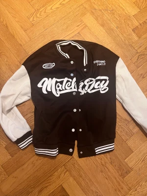 Brun och vit bomberjacka Match Day - Snygg svart och vit bomberjacka från Match Day med stora tryck på framsidan och coola detaljer som randiga muddar och knappar. Jackan har långärmade vita ärmar och svart kropp, samt texten 'Last Call' och 'Everything I Get It' på bröstet.