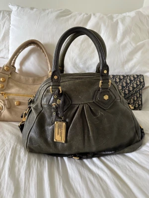 Snygg Q Classic Aidan bag! - Perfekta skolväskan! Stilren och rymlig mörkgrön Marc by Marc Jacobs Classic Q Aidan i äkta läder med guldfärgade detaljer och dubbla handtag. Långa bandet finns. Den höga och eleganta formen gör att den rymmer en 13” MacBook tillsammans med allt du behöver för skola, jobb eller fritid. Finns slitage på lädret som syns på bilderna. Väskan har dragkedja upptill och praktiska innerfack för smidig organisering. Kan bäras i handen eller på axeln. En eftertraktad Q-Classic väska. Mått ca 26×36×14 cm🤍