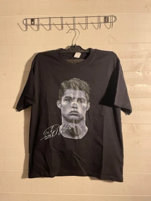 Svart t-shirt med tryck och signatur - Cool svart t-shirt med stort tryck av ett ansikte och vit signatur på framsidan. Baksidan har en stor grafisk design med texten 'CUT ANYTHING YOURS'. Klassisk passform och rund hals. Perfekt för dig som gillar streetwear vibes.